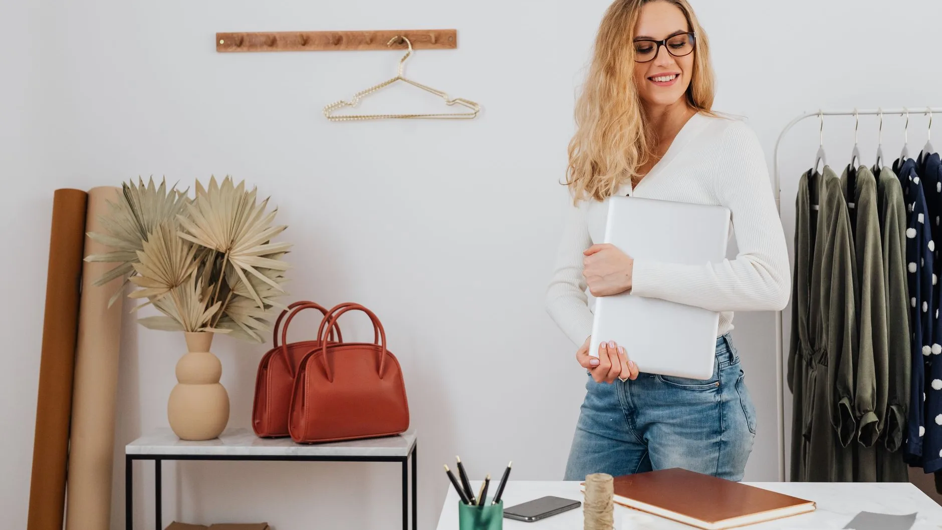 A fashionable woman holding a laptop stands in a chic boutique setting with clothes and accessories.